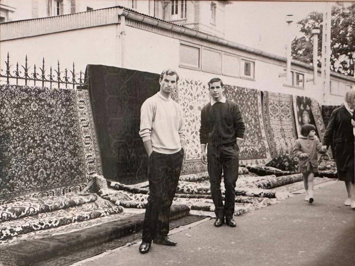 Photographie ancienne : deux jeunes hommes devant un mur de tapis présentés dans la rue